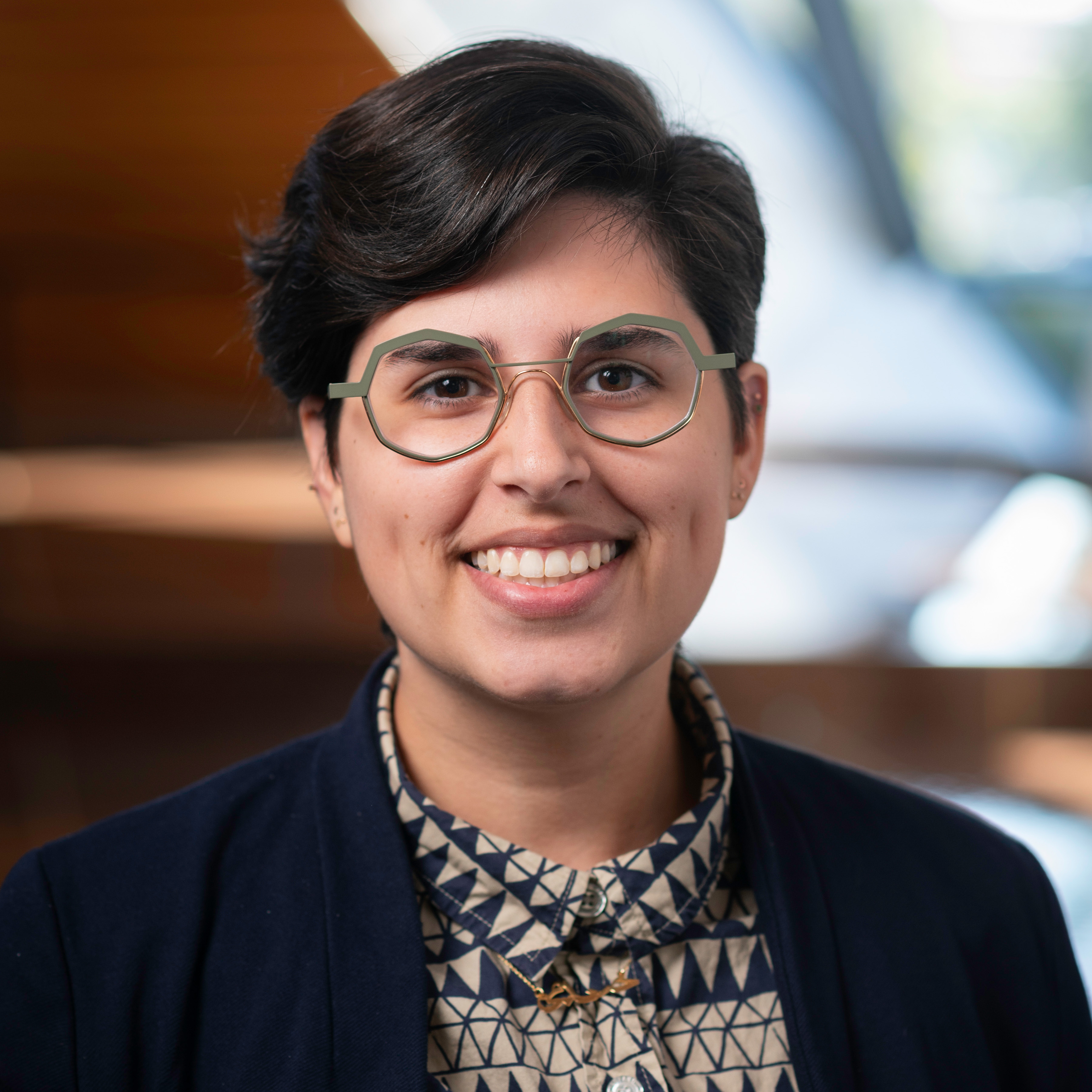 A person with tan skin and dark hair wearing a patterned dress shirt, a black blazer, and silver glasses