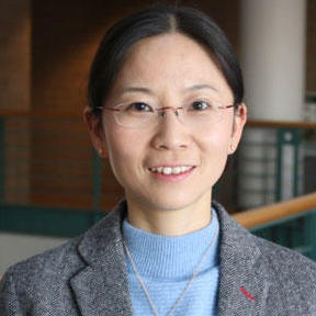 A woman with light skin and dark hair wearing a blue sweater, grey blazer, a silver necklace, and clear glasses. 