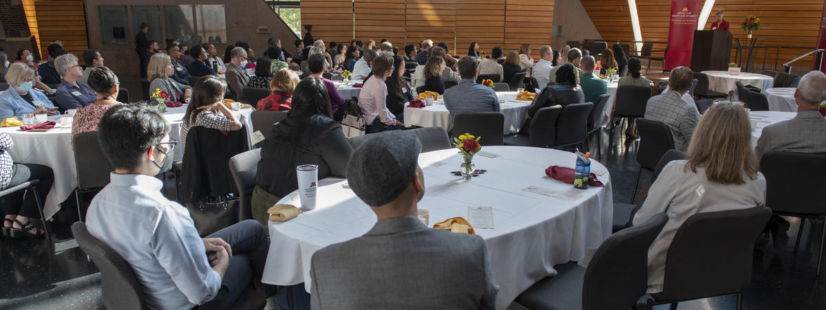 Photo from the Fall Welcome Breakfast 2023. It is taken from the back of the room, and all participants are paying attention to the speaker at the podium.